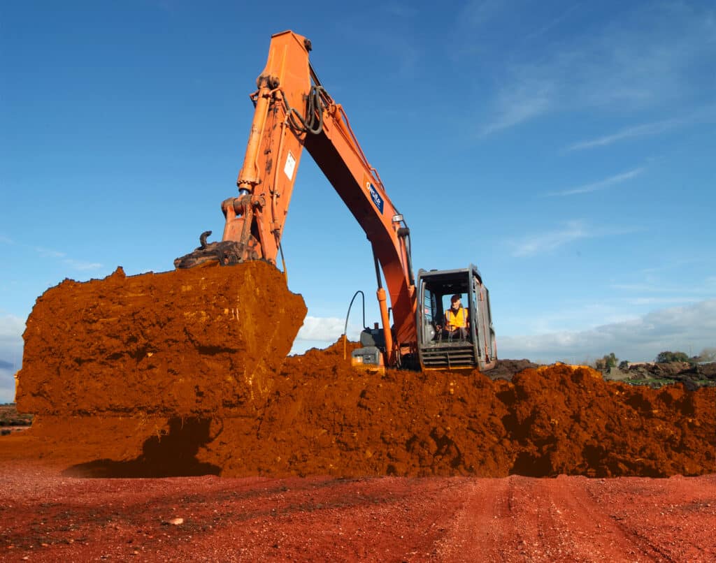 A man operates a large rented excavator, scooping red soil