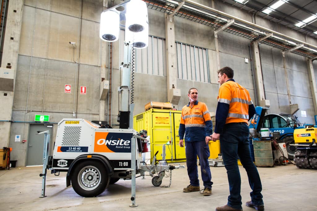 Two Onsite staff in hi-vis jackets standing next to some equipment hire and talking to each other in an industrial setting