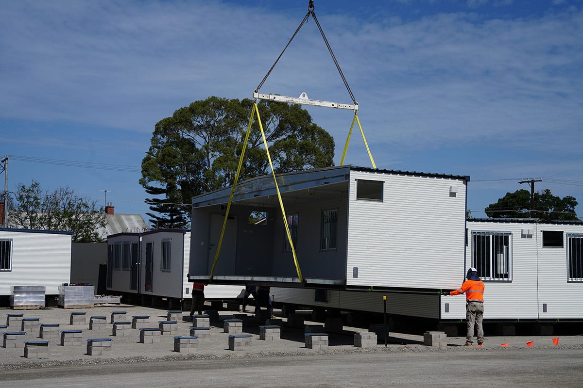 A prefabricated Onsite building module being lifted into place by a crane at work site, with workers guiding the unit onto concrete footings.