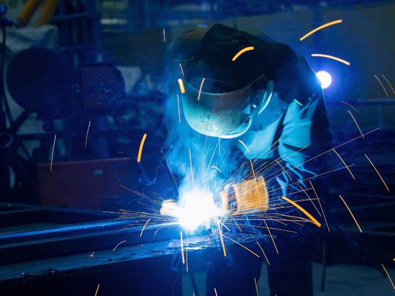 Welder in protective helmet and gloves MIG welding steel beams, with bright arc and orange sparks flying in an industrial workshop